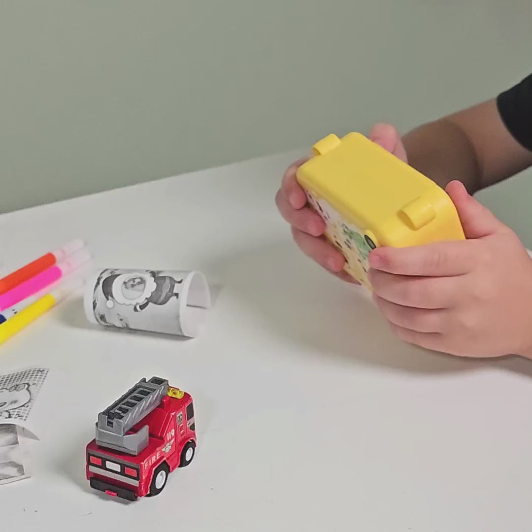 Boy using the Instant Print Kids Digital Camera to take a picture of a small red fire truck at a table. Colorful markers are in the background, used for coloring printed photos. The camera screen displays its features before the boy excitedly photographs a puppy and instantly prints the image. Later, he explores the camera’s fun built-in frames, discovering many creative options.